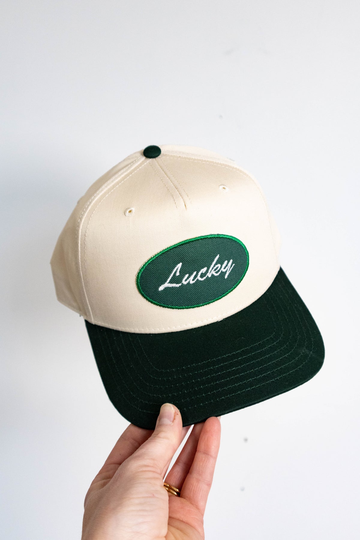 Beige and black cap with 'Lucky' logo held by a hand on a white background